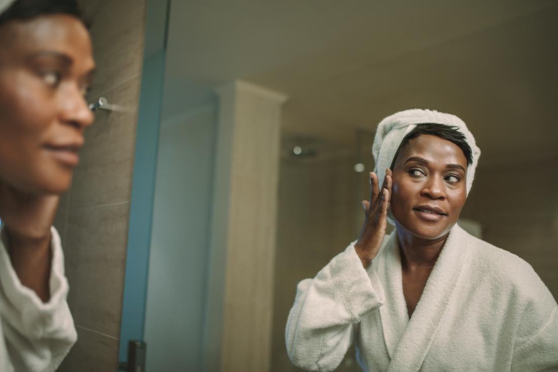 Woman in front of bathroom mirror applying CBD oil or moisturizer to her face to treat acne