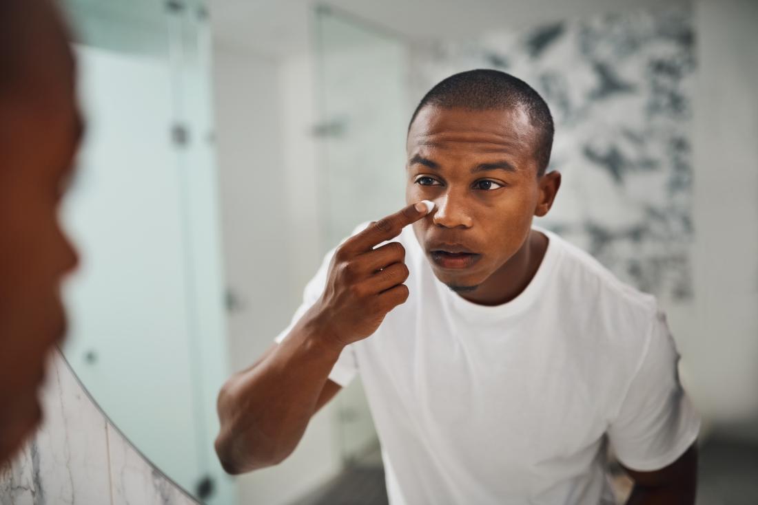 Black or african american man looking in bathroom mirror applying cream or lotion to skin under eyes.