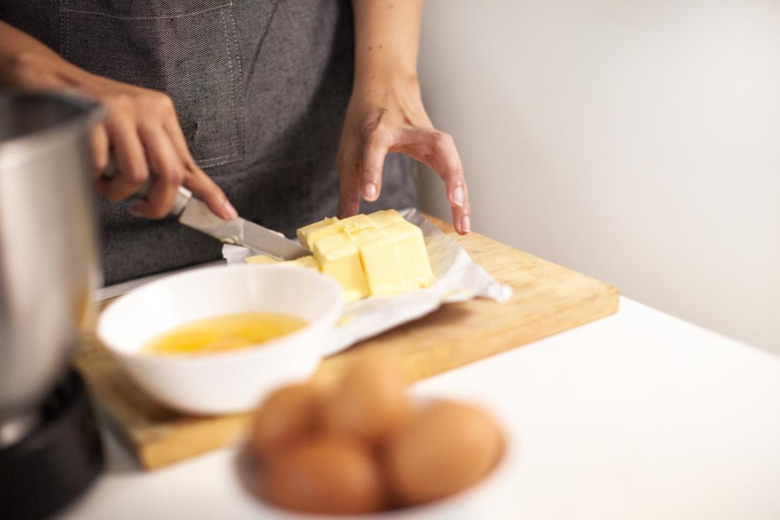 Person cutting up butter on wooden chopping board in kitchen with oil and eggs in the foreground.