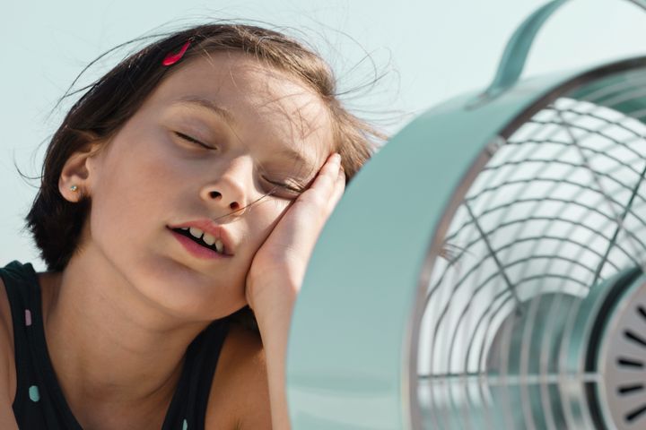 Overheated little child is sitting in front of electric fan trying to cool down.